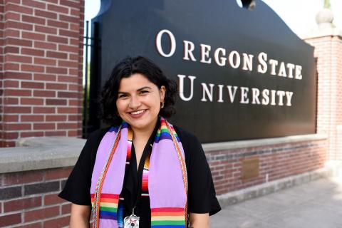 photo of sienna kaske wearing a lavendar and rainbow graduation stole, sarapé graduation stole, orange, gray and orange and blue cords with a black jumpsuit smiling in front of an Oregon State Unviersity black sign